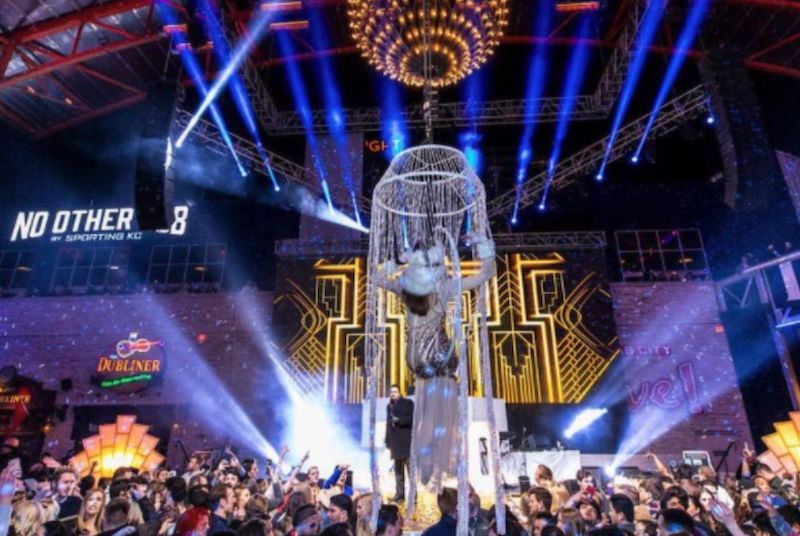 A lighted ball hangs over a glittery-dressed acrobat sitting in a hoop above the crowd as an emcee looks on.