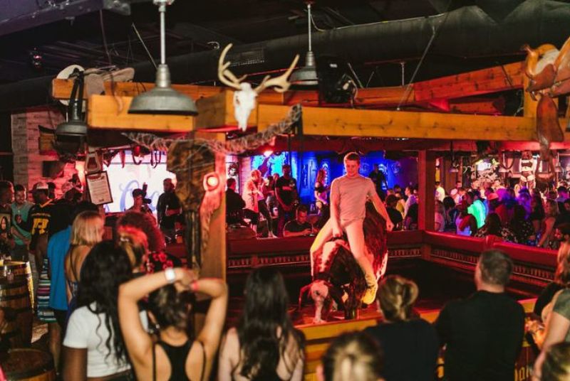 A man leans back astride a mechanical bull that holds its head low and hindquarters very high while an excited crowd watches.