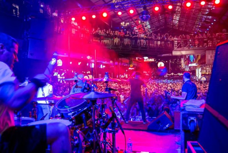 Looking out from behind a drummer mid-beat, a band is bathed in red light as the singer faces an excited wall-to-wall crowd!