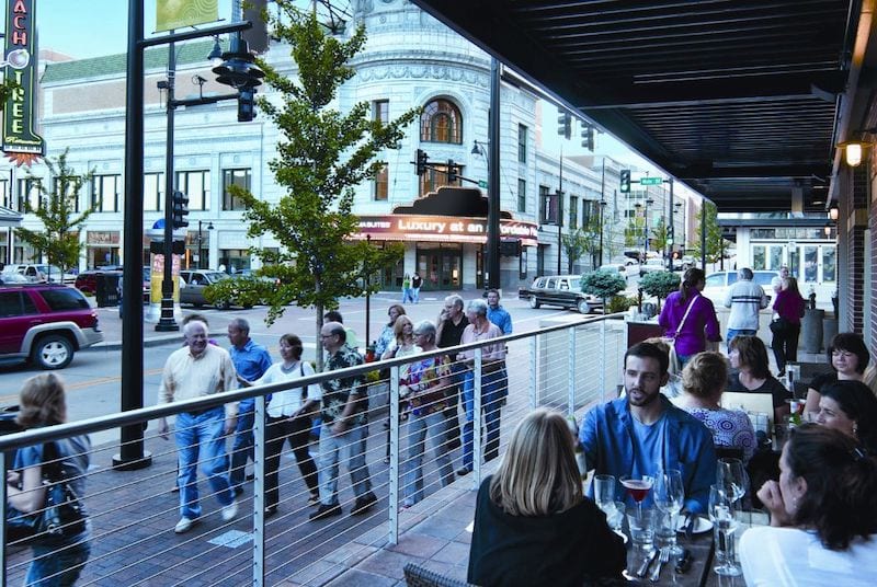 Two tables of diners enjoy drinks on a lovely patio and people-watch as groups walk by on the sidewalk a few feet away.