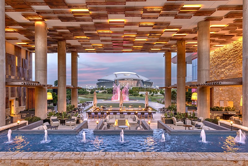 An outdoor bar and plush seating areas surrounded by fountains overlook a sunset and crescent moon behind AT&T stadium.