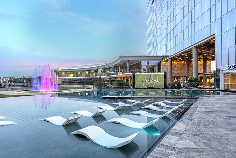 An inviting outdoor infinity pool overlooks a purple and pink-lit fountain as an NFL game plays on a large LED screen nearby.