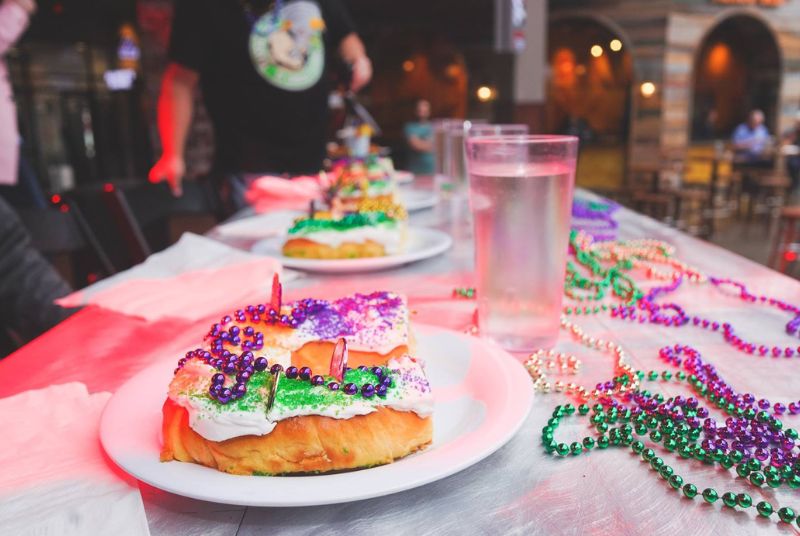 Slices of king cake and glasses of water line a table decorated with Mardi Gras beads.