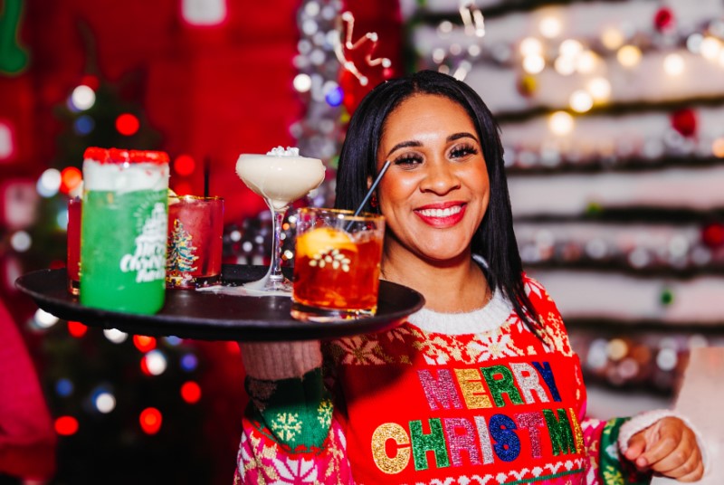 Smiling waitress wearing "Merry Christmas" sweater, holding a tray of holiday-themed drinks.