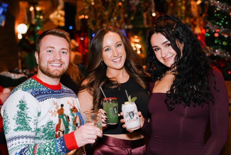 Man in a holiday sweater with two women toasting holiday-themed cocktails.