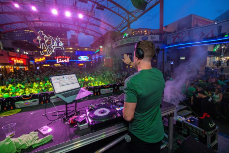 A DJ dressed in green with a shamrock neck tattoo looks out over a large dancing crowd at Kansas City Live!