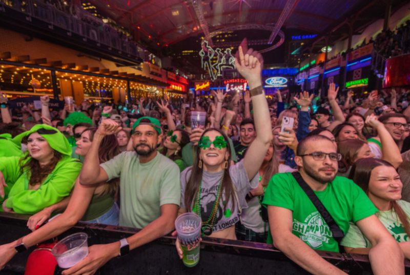 A smiling woman with shamrock glasses puts her hand in the air as an enthusiastic crowd dances behind her.