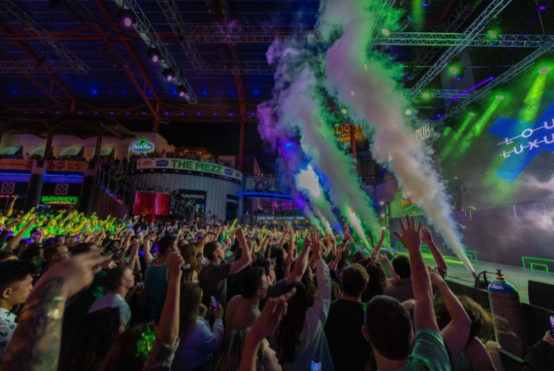 Green spotlights shine on plumes of smoke at the front of a stage as an excited crowd thrusts their arms up in anticipation!