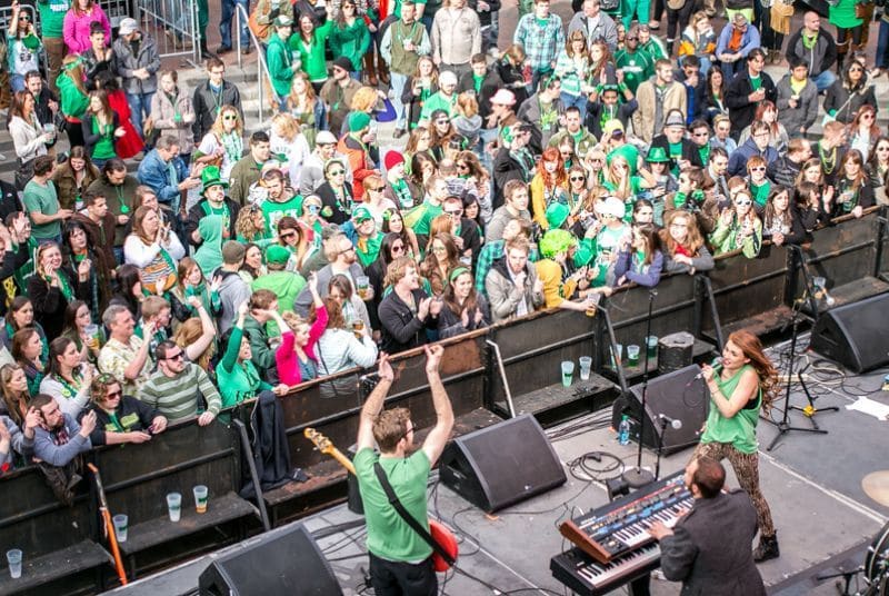 A smiling St. Patrick's Day crowd watches as a guitarist, female singer, and keyboard player perform.