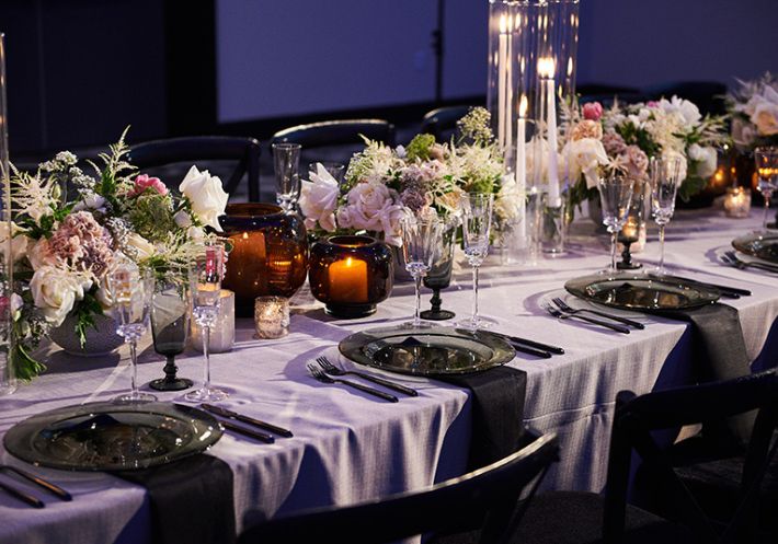 An elegant lavender table holds pale pink & purple floral arrangements bordered by candles of all sizes and place settings.