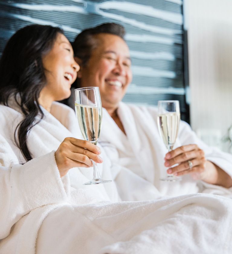 Middle aged Asian couple sitting in bath robes on a bed with glasses of champagne