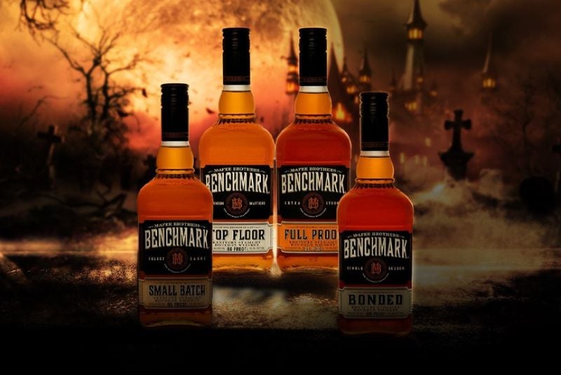 Four different bottles of Benchmark brand liquor on an orange and black, spooky, forest background.