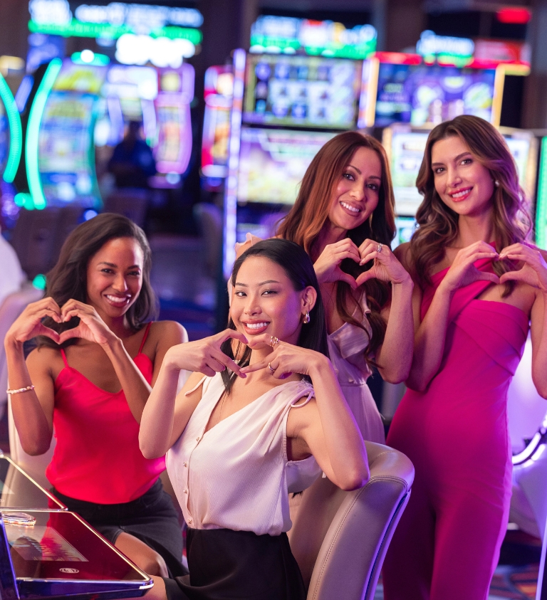 4 women holding up hearts with their hands standing by slot machines