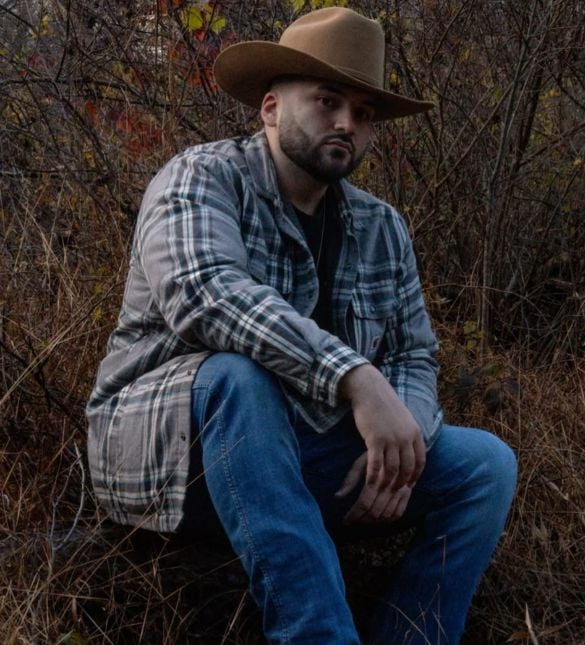 A country musician in a plaid shirt and cowboy hat seated outdoors in winter brush.