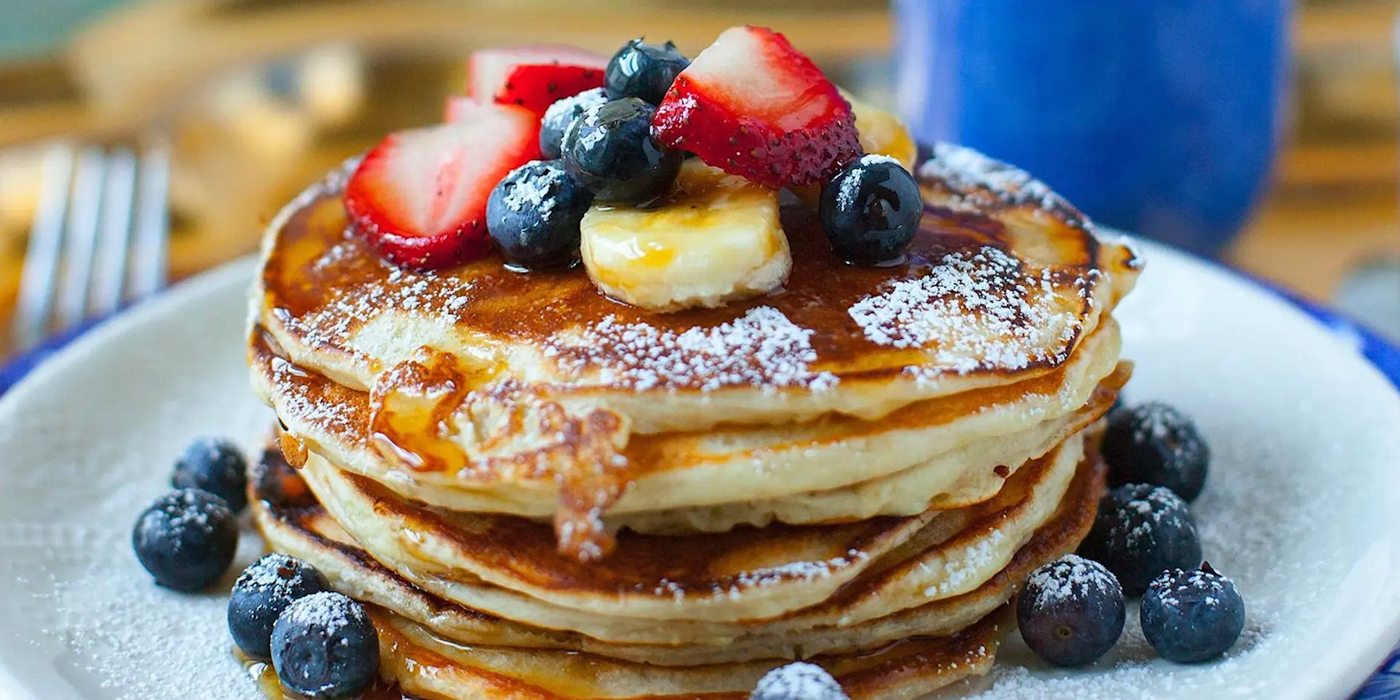 Stack of fluffy pancakes topped with fresh berries, banana slices, powdered sugar, and syrup on a white plate.