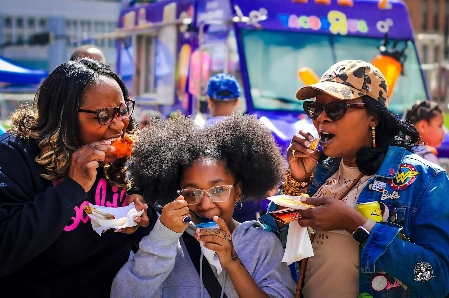 Three people enjoying food together outdoors in front of colorful food trucks at a festival.