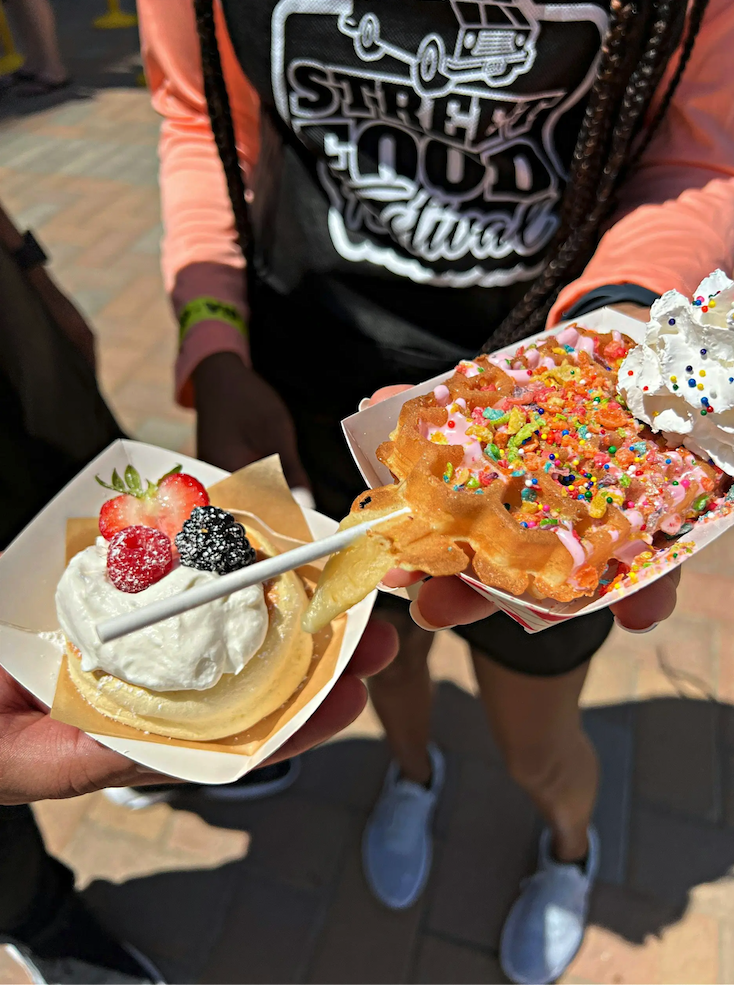 Two dessert waffles topped with whipped cream, fruit, and colorful cereal toppings being held at a street food festival.