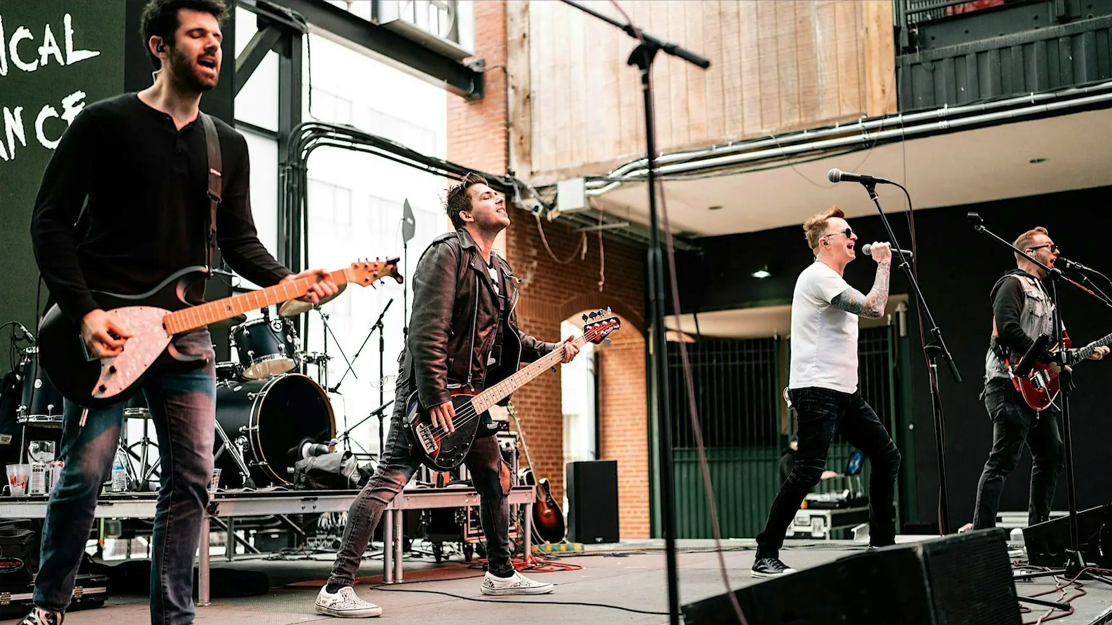 Rock band performing live on an outdoor stage with guitars, drums, and microphones.