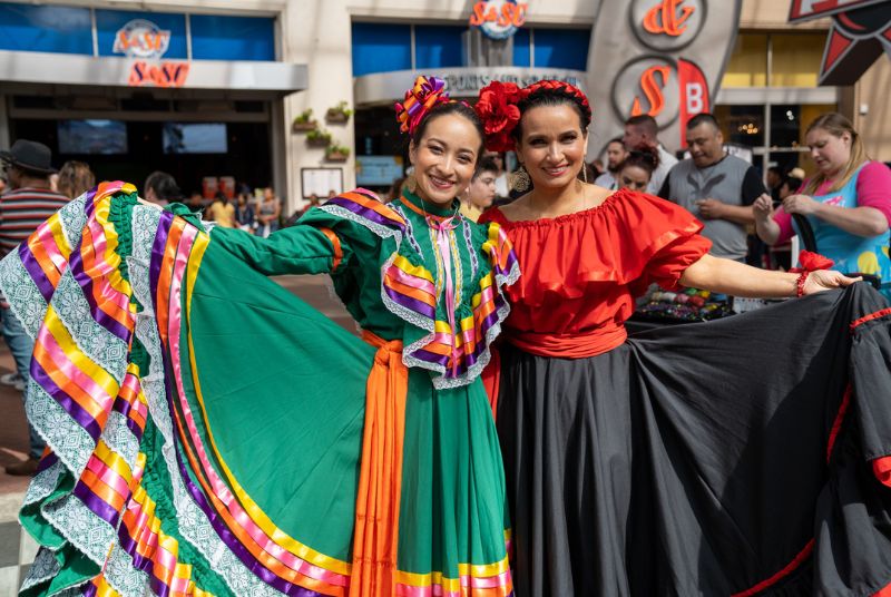 Two Cinco de Mayo dancers wear traditional dresses, one green with multi-colored ribbons and lace, the other red and black.