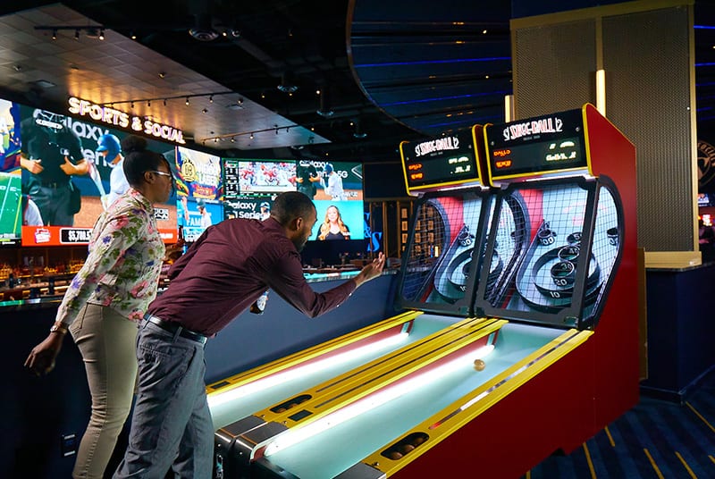 A man takes his turn at Skee-Ball as his female companion looks on. A media wall shows live pro sports games behind them.