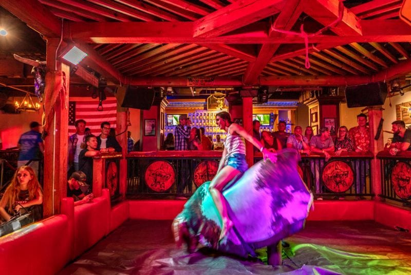 A woman in a tank top and jean shorts rides a mechanical bull at PBR as the crowd watches.