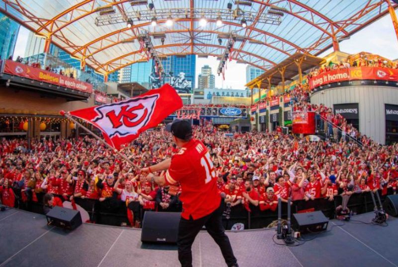 On stage a man waves a Kansas City Chiefs flag as a packed crowd cheers during the Super Bowl watch party at Kansas City Live!