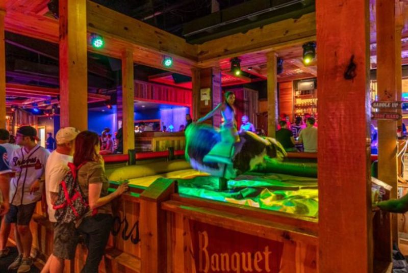 A woman in a tank top rides a fast-moving mechanical bull at PBR as onlookers watch.