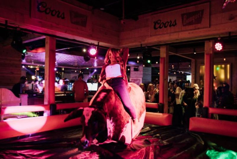 A smiling woman leans back astride a mechanical bull that holds its head low and hindquarters high while a crowd watches.