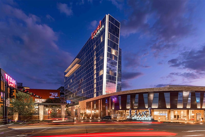 A pink and purple sunset is reflected in the side windows of the Live! by Loews Hotel as car lights whiz by.