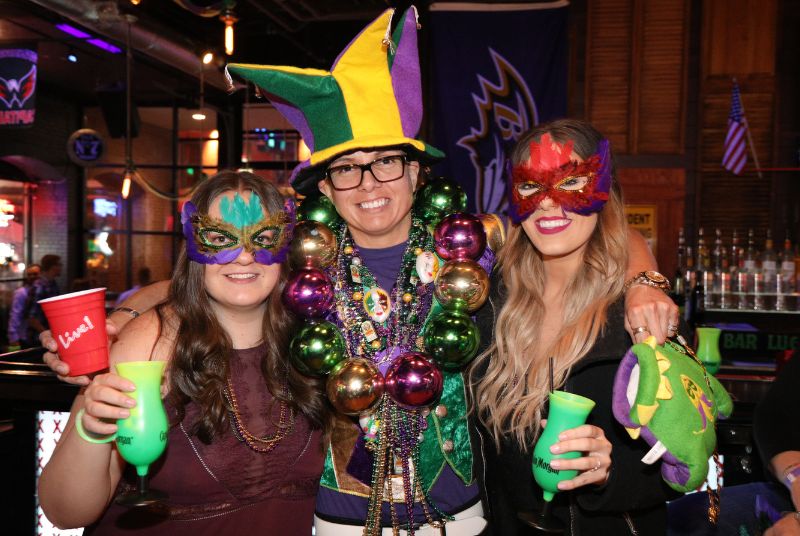 A jester wearing a large number of beads and a massive necklace poses with two women in feathered Mardi Gras masks.
