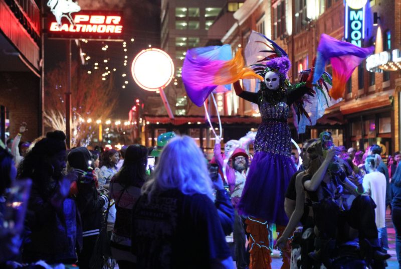 A performer in a white full-face mask with a beaded gown and rainbow-feathered wings waves colorful scarves above the crowd.