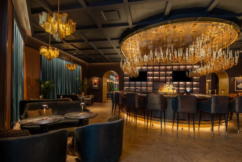 Dim lit lounge with tables and chairs and a bar area. Velvet blue chairs and curtains, lots of hanging light fixtures.