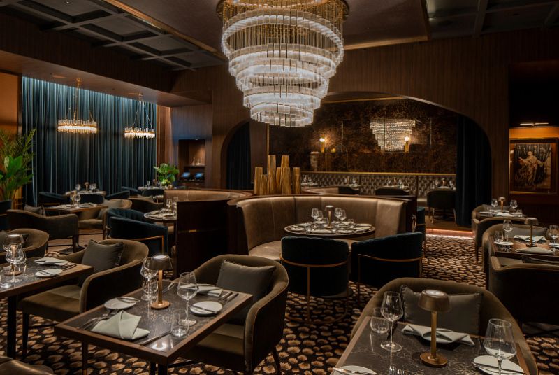 Large dining room with booths and tables. Some tables have velvet blue, large chairs. Velvet blue curtains hang on the far side of the room. A large chandelier hangs in the middle of the room.