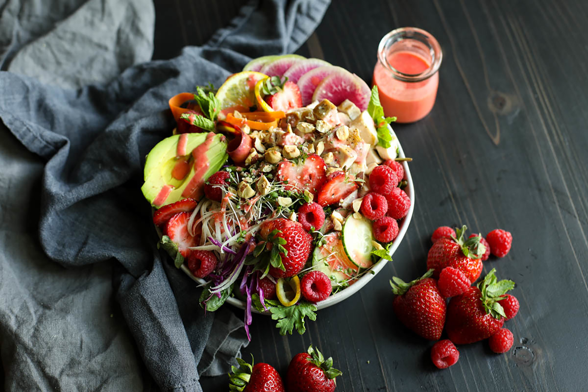 Strawberry protein salad topped with berries, avocado, nuts, radish. White bowl against a black and gray background with a dark linen napkin. 