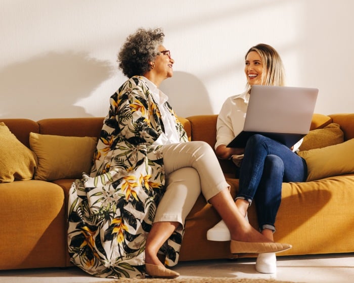 Photo of two women sitting on a couch, smiling, while looking at a laptop