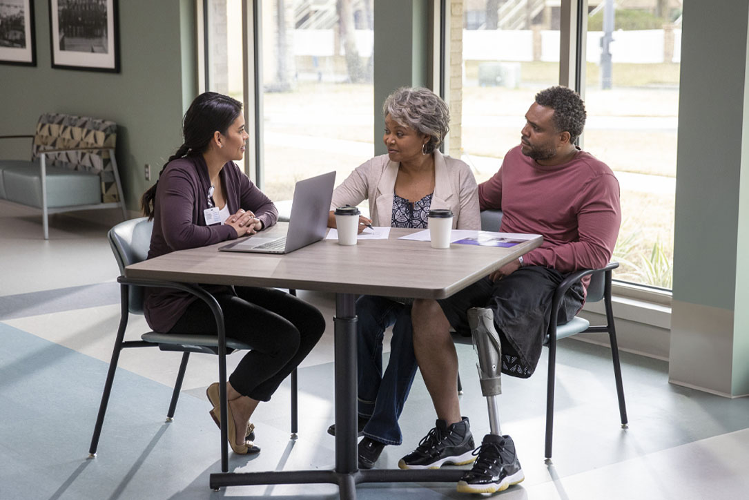 Three people sit at a table in a bright room, engaged in discussion. A laptop and coffee cups are on the table. The atmosphere is collaborative.