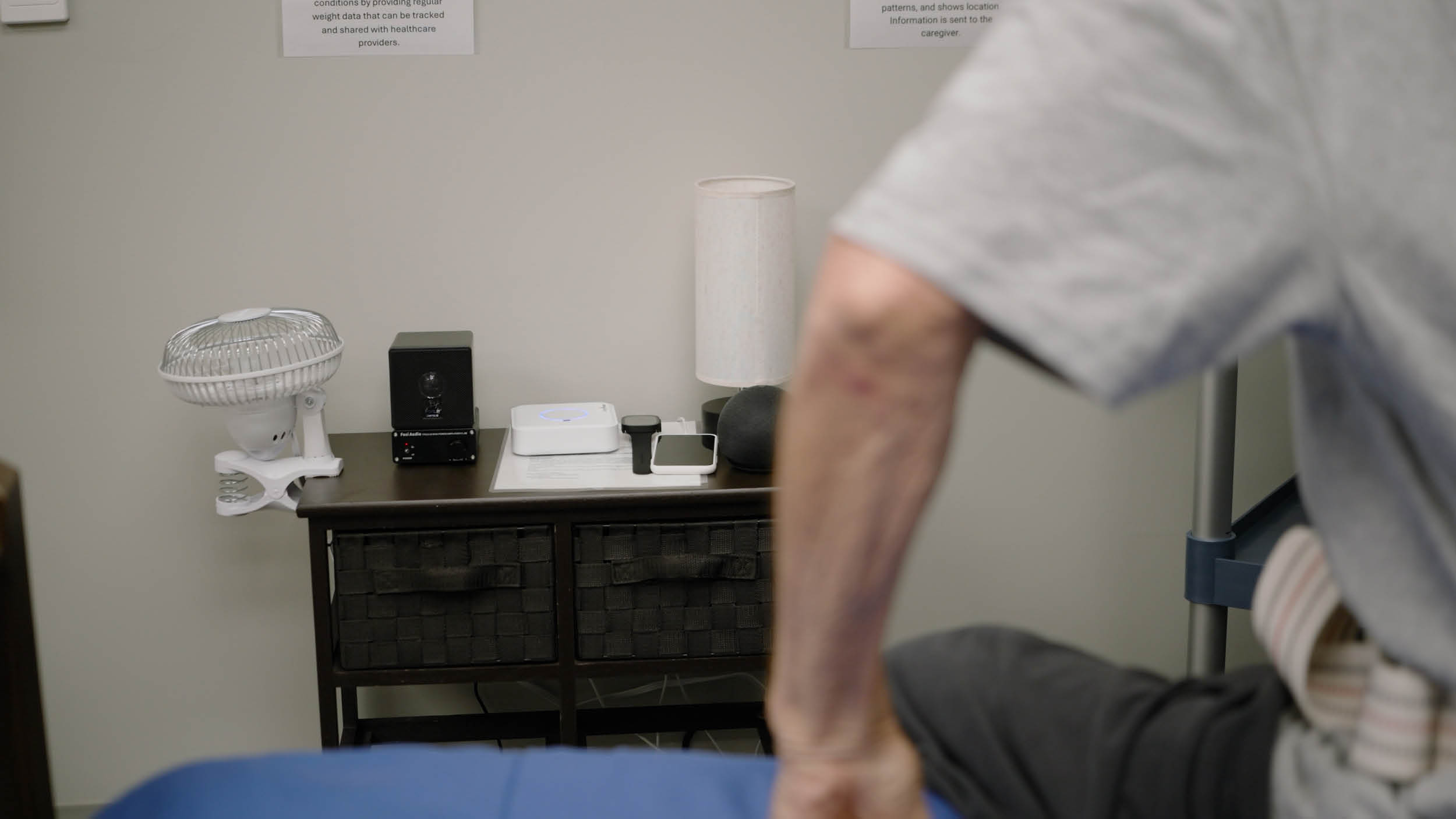 A person with a support belt sits on a massage table, facing a table with a fan, speaker, lamp, and other items, creating a clinical setting.