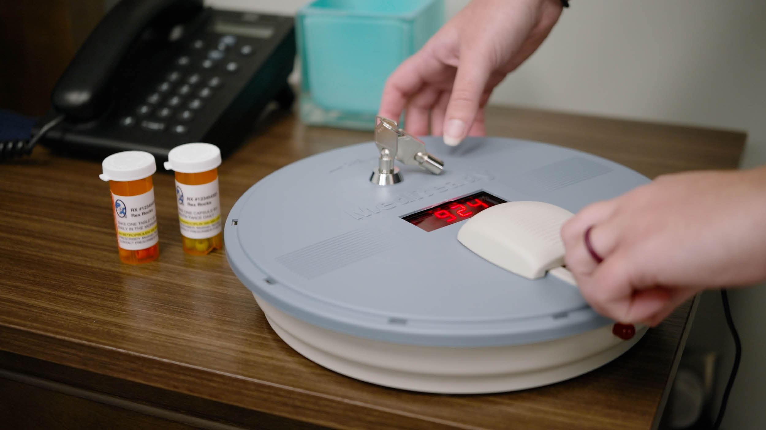 Hands interact with a gray timer lockbox on a desk displaying 9:24. Nearby are two prescription bottles and a cordless phone, suggesting a secure medication setup.