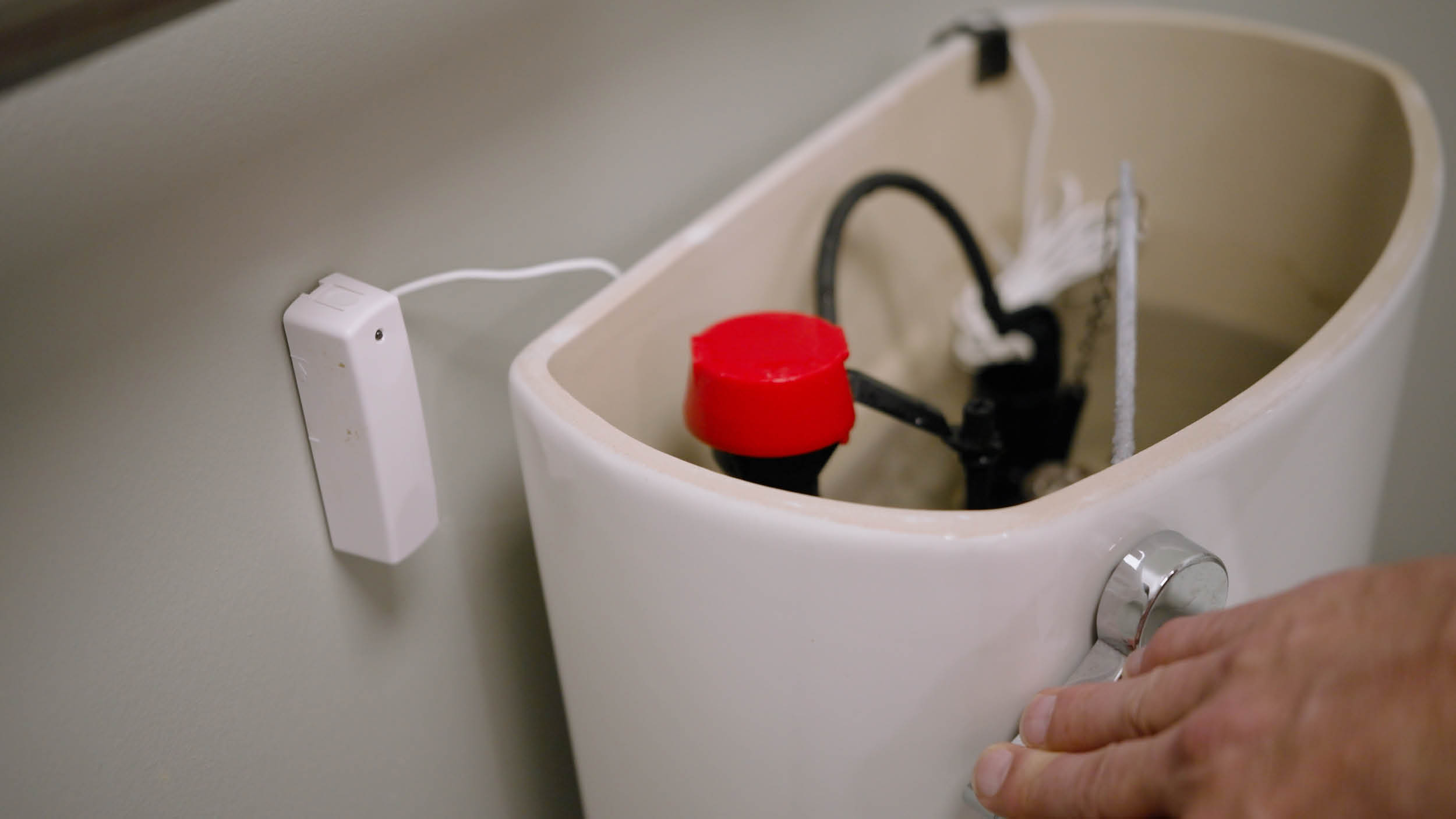 Toilet tank lid removed, showing internal parts including a red float. A sensor is attached nearby. A hand reaches for the flush handle, indicating interaction.
