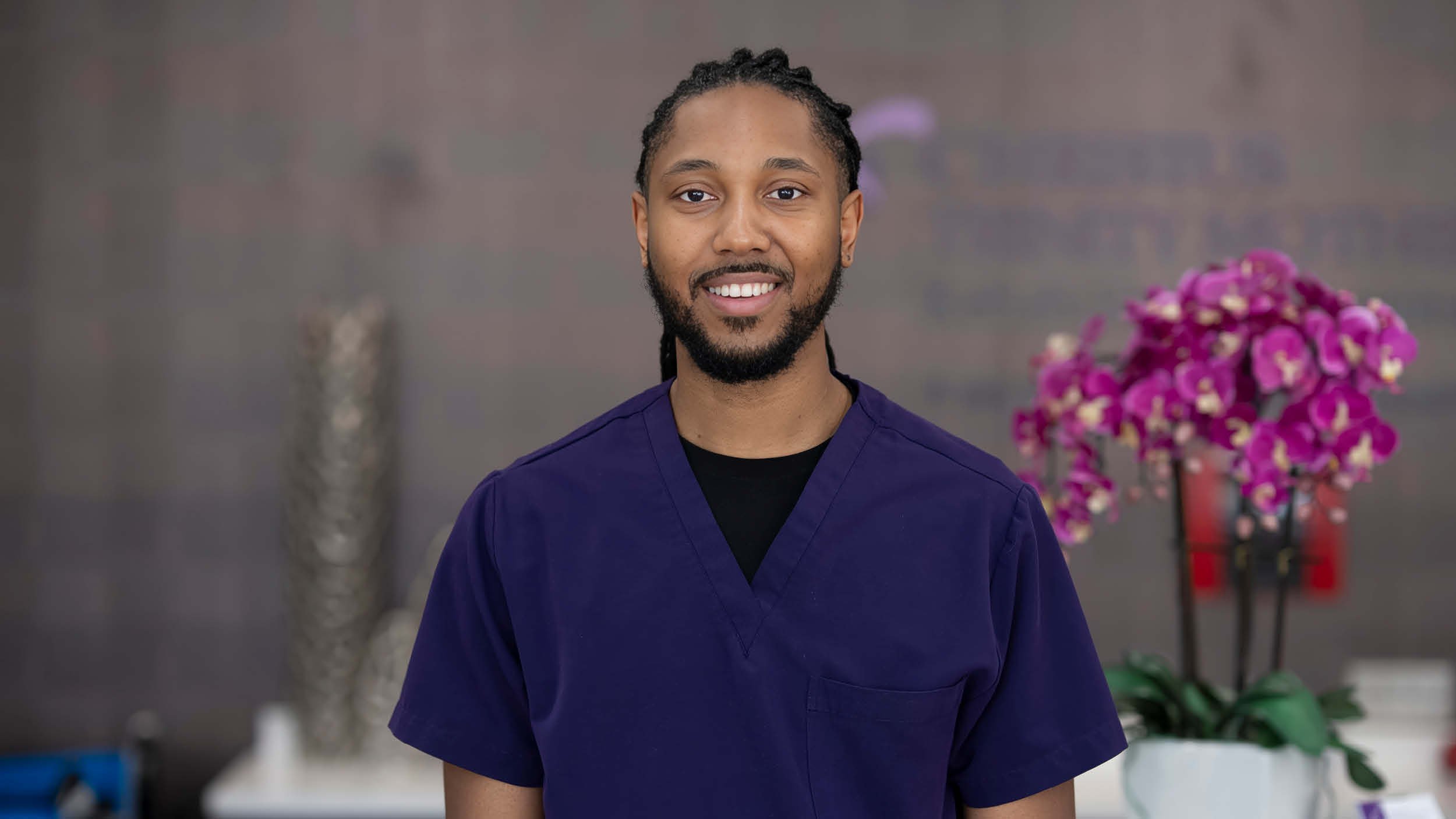 Rehabilitation hospital employee Trevor Johnson recognized as OEAA Southwest winner, pictured in dark blue scrubs. Behind him, bright magenta orchids add a vibrant touch to the neutral setting.