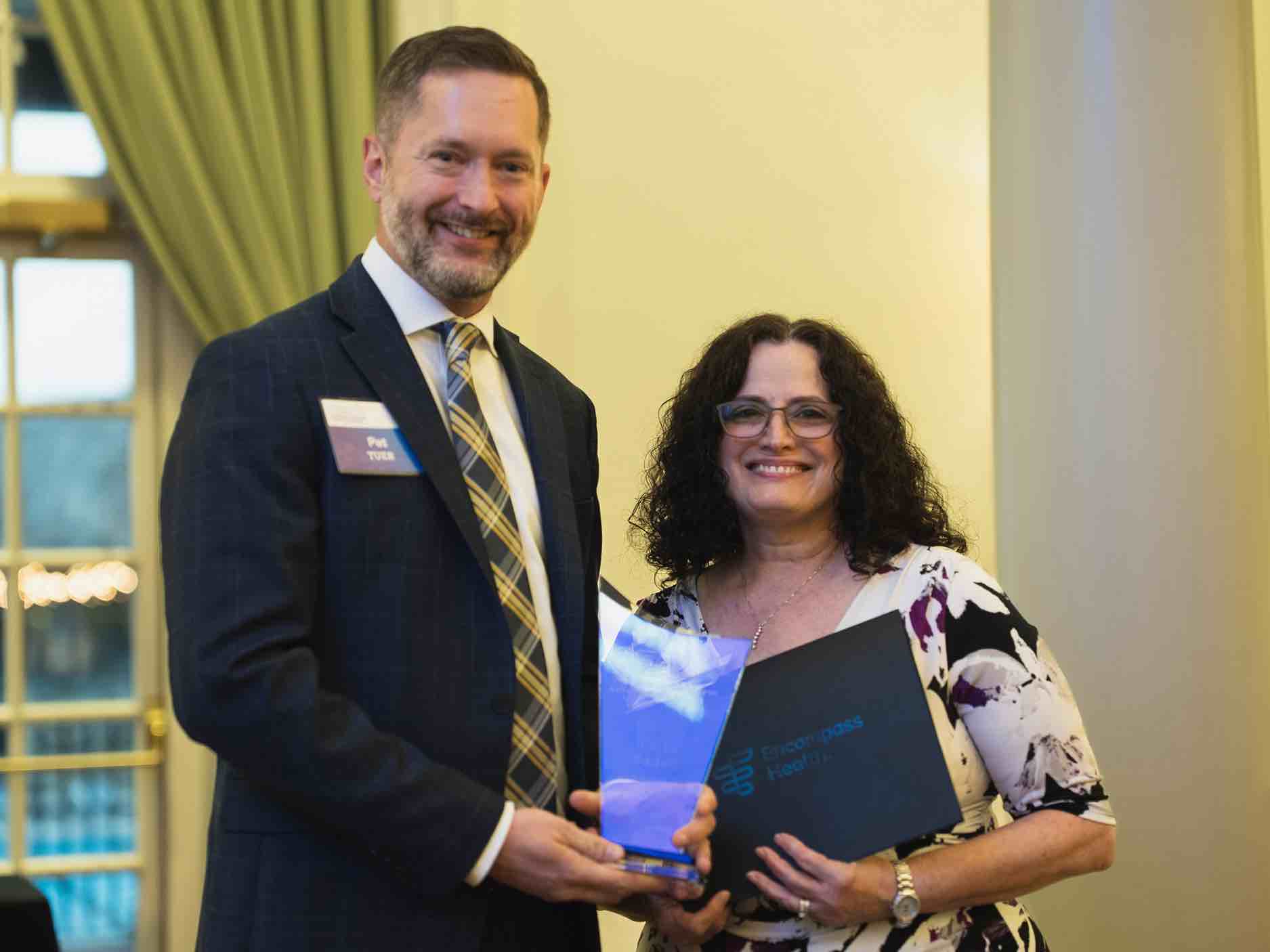 Evangelina Badell stands at an OEAA awards presentation, holding a recognition plaque alongside a colleague, representing leadership and excellence in rehabilitation health care achievement and service.
