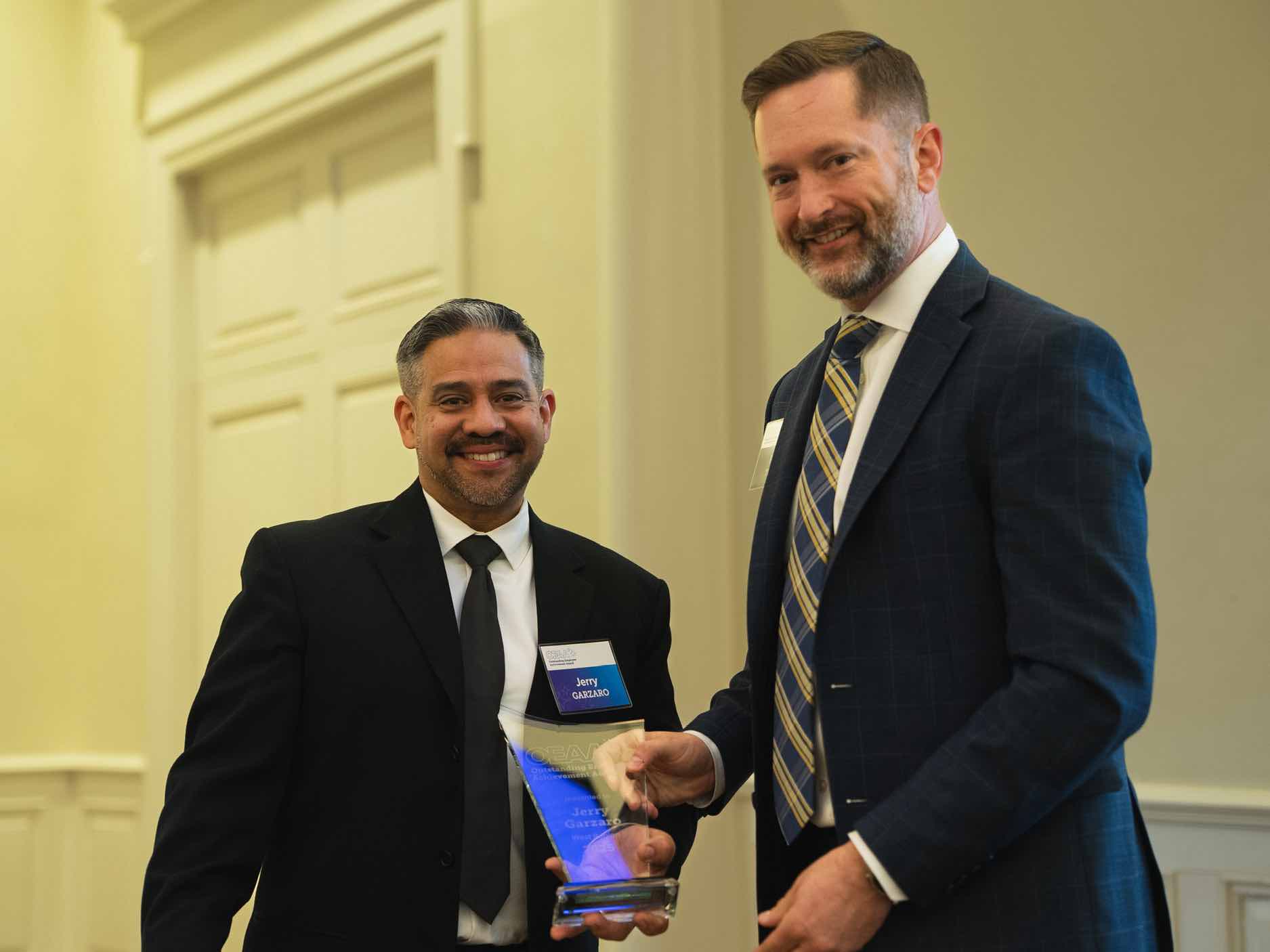 Jerry Garzaro stands at an OEAA awards presentation, holding a recognition plaque alongside a colleague, representing leadership and excellence in rehabilitation health care achievement and service.