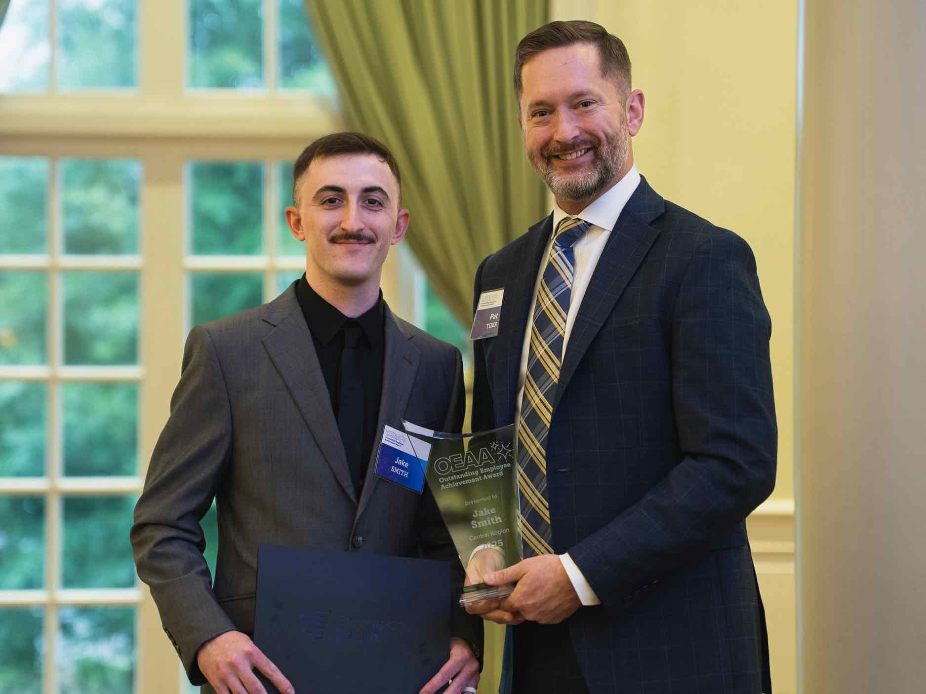 Jacob Smith stands at an OEAA awards presentation, holding a recognition plaque alongside a colleague, representing leadership and excellence in rehabilitation health care achievement and service.