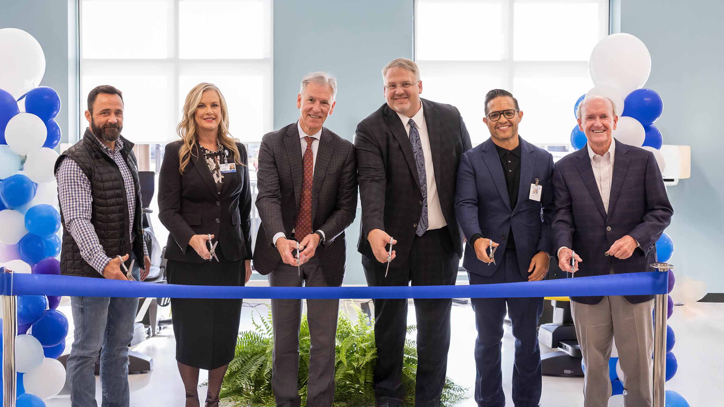 Seven people smiling and holding scissors stand behind a blue ribbon at a ceremony. 