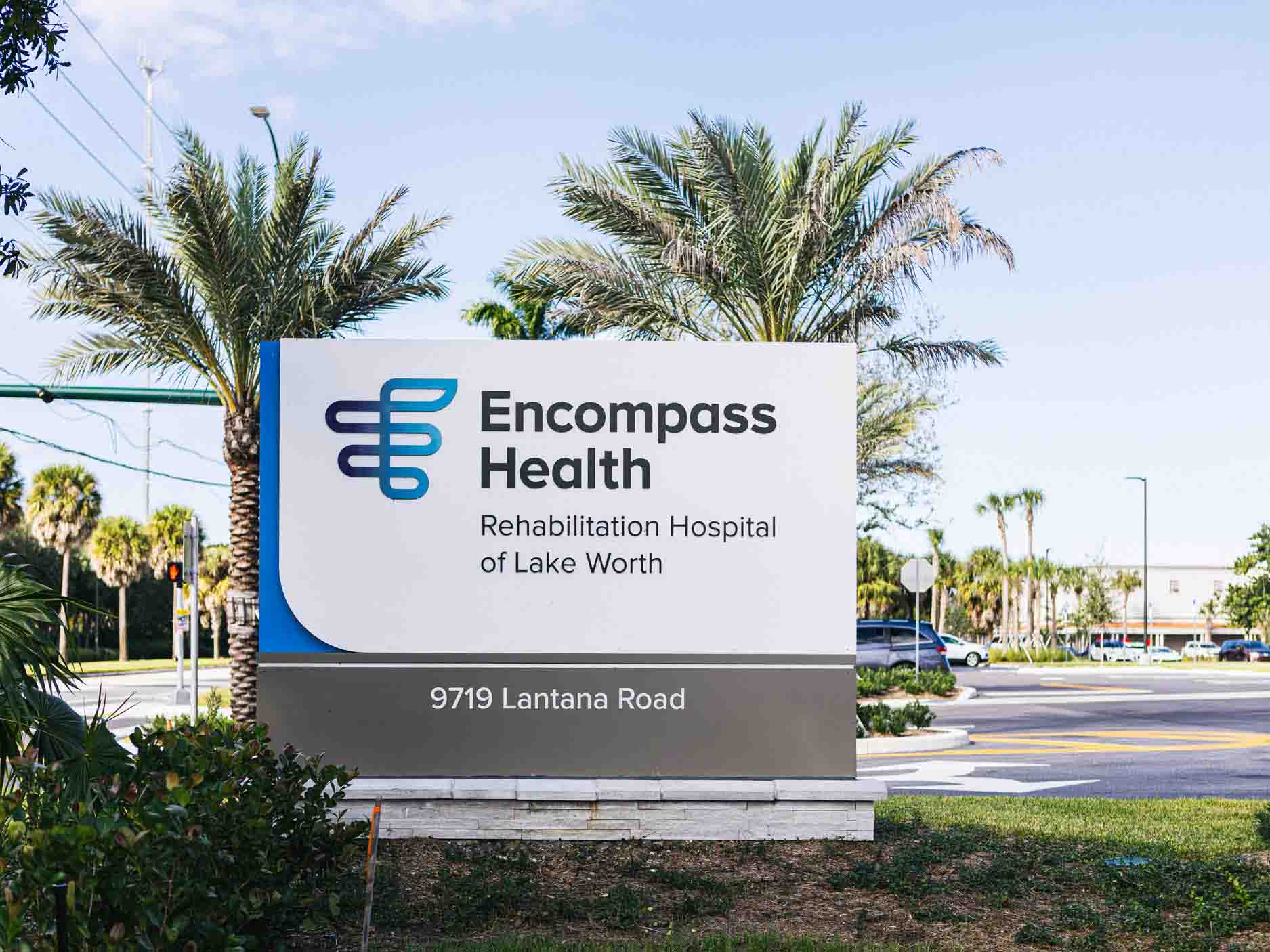 Sign for Encompass Health Rehabilitation Hospital of Lake Worth at 9719 Lantana Road. Palm trees and a street are visible in the background.