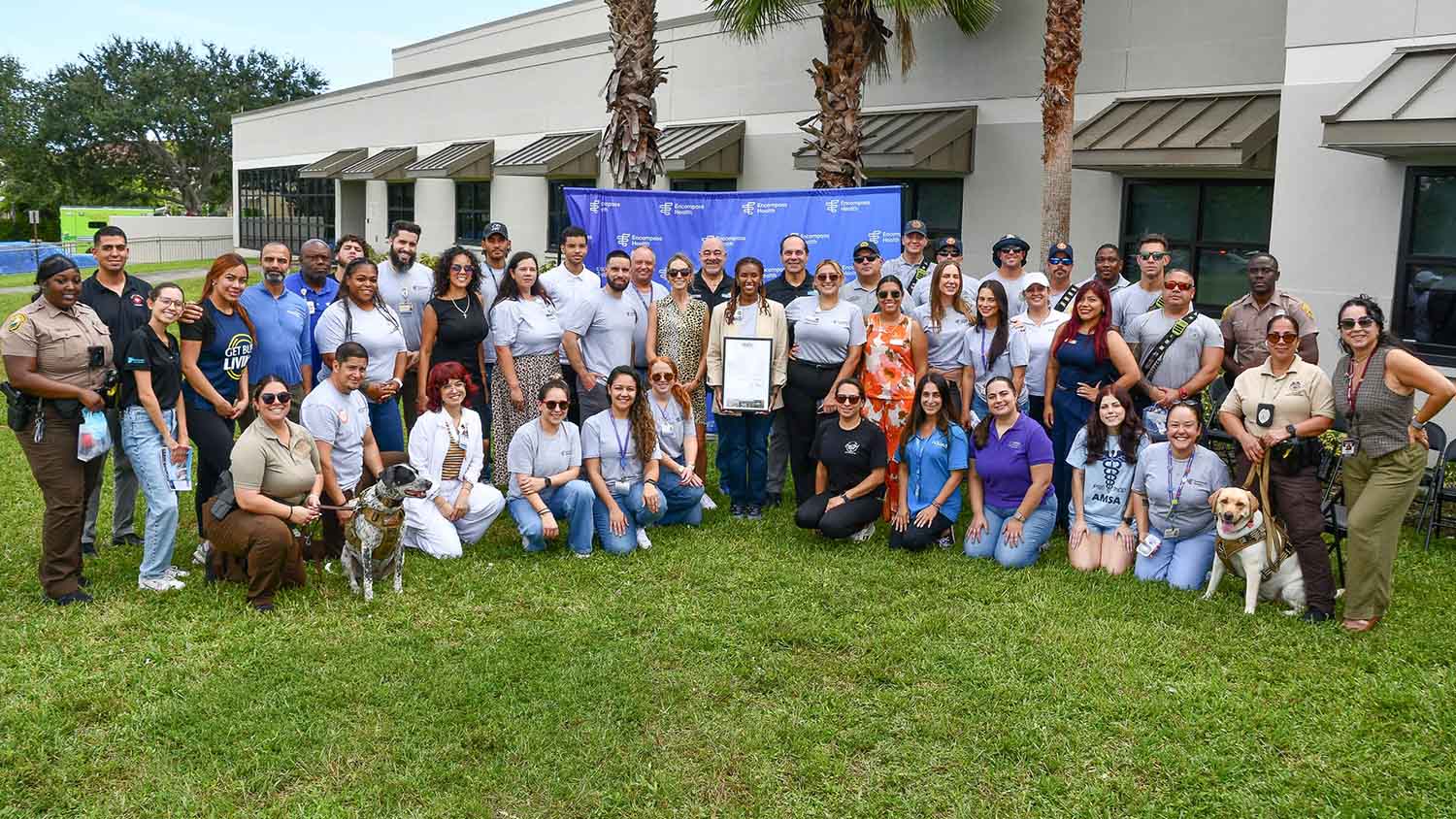 A diverse group of people, including animal handlers with dogs, gathered outdoors in front of a building, smiling and posing for a group photo.