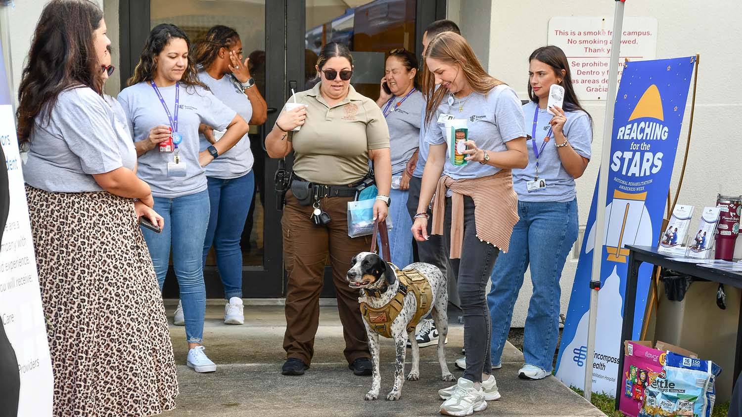 A group of smiling people, some in uniform, gather outside a building with a therapy dog wearing a vest. A 