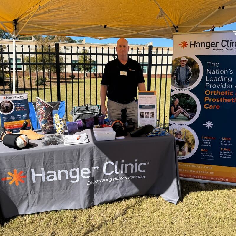 A man stands under a yellow tent behind a table displaying prosthetic devices with a Hanger Clinic banner. The setting is outdoors with a fenced building in the background.