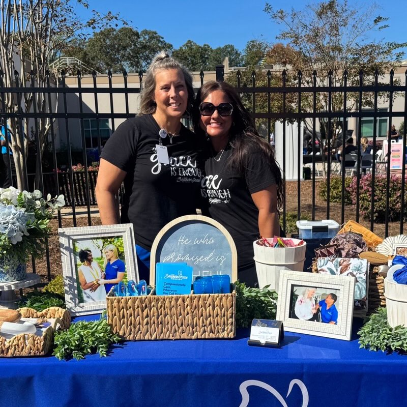 Two women smiling behind a table with a blue cloth, featuring photos, and gift baskets. Sunny outdoor setting.