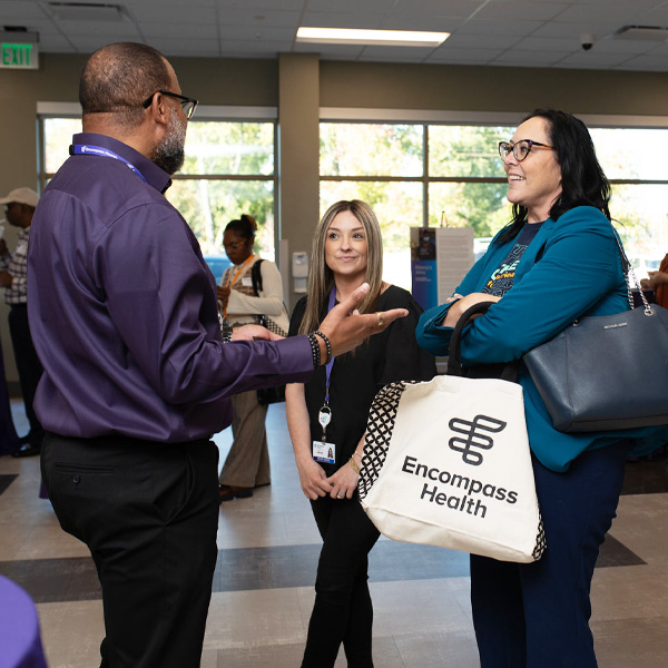 Encompass Health Toledo employees converse in a brightly lit room. One person, wearing a purple shirt, gestures while facing two women, one holding an “Encompass Health” tote.